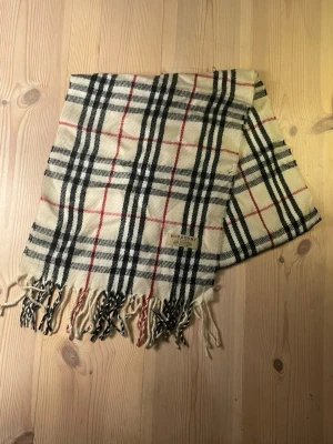 Burberry halsduk i klassiskt rutmönster - Snygg halsduk från Burberry i beige med det ikoniska rutmönstret i svart, vitt och rött. Halsduken har fransar i ändarna och är tillverkad i mjukt material, perfekt för att lyfta vilken outfit som helst. Pris kan diskuteras 