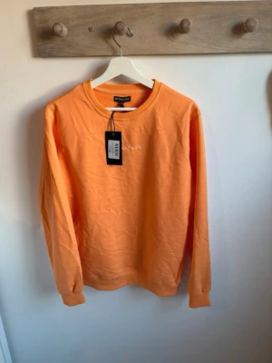 Orange sweatshirt från On That Ass - Snygg orange sweatshirt  med rund halsringning och diskret broderad text på bröstet. Tröjan har långa ärmar och ribbade muddar vid ärmslut och nederkant. Perfekt för en avslappnad och trendig look.