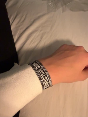 Dior armband - Säljer ett svart och vitt tygarmband från Christian Dior med texten 'CHRISTIAN DIOR J'ADIOR'. Armbandet har justerbar knytning och en stilren, trendig design som passar perfekt till streetwear eller som en snygg detalj till din outfit.