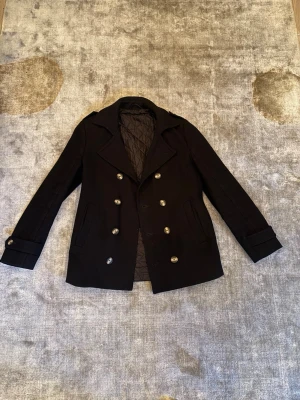 Svart dubbelknäppt rock - Stilren svart skepparkavaj med dubbelknäppning och stora silvriga knappar. Jackan har klassisk krage, två fickor framtill och quiltat foder på insidan. Perfekt för dig som vill ha en tidlös och snygg look under kallare dagar.