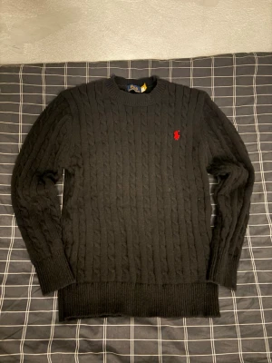 Svart stickad tröja från Polo Ralph Lauren - Svart kabelstickad tröja från Polo Ralph Lauren med klassisk rund hals och röd broderad logga på bröstet. Långärmad och i mjukt stickat material, perfekt för kyligare dagar. Snygg och tidlös modell som funkar till allt. Använd 1 gånger. Är i nyskick, hör av dig om du har några frågor.