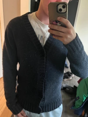 Mörkblå stickad kofta med knappar - Säljer en mörkblå stickad kofta med v-ringning och stora knappar framtill. Koftan har långa ärmar och breda ribbade muddar vid ärmslut och nederkant. Perfekt att slänga på sig när det är kyligt ute.