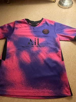 PSG matchtröja rosa/lila - Säljer en PSG matchtröja i en riktigt fet rosa och lila färgkombination med svart logga och detaljer. Tröjan har ett unikt mönster och V-ringning. Perfekt för dig som vill sticka ut på planen eller på stan.