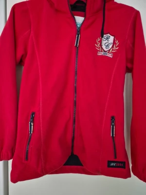 Fleecejacka - Röd fleecejacka med huva från Mywear Active. Jackan har dragkedja framtill, två fickor med dragkedjor och broderad detalj på bröstet. Perfekt för kyliga dagar och enkel att slänga på sig. Långärmad och mjuk i materialet. Några fläckar.