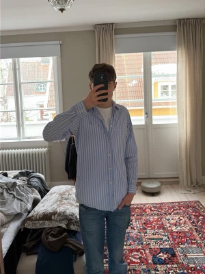 Randig skjorta från Ralph Lauren - Säljer en klassisk skjorta från Ralph Lauren med blå och vita ränder samt inslag av ljusrosa. Skjortan har lång ärm, knappar framtill och ikonisk broderad logga på bröstet. och har en relaxed fit.