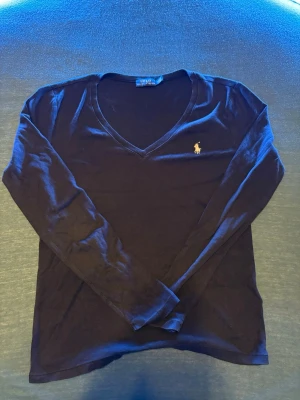 Svart långärmad tröja Polo Ralph Lauren - Klassisk svart långärmad tröja från Polo Ralph Lauren med v-ringning och den ikoniska broderade loggan på bröstet. Tillverkad i mjuk bomull som känns skön mot huden. Perfekt till jeans eller kjol för en clean och stilren look. Finns någon fläck men går att tvättas 