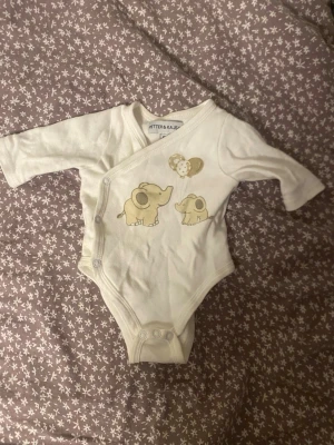 Vit body med elefanter Petter & Kajsa - Supersöt vit body från Petter & Kajsa i storlek 50. Tryck framtill med två beige elefanter och ballonger i mjuka toner. Omlottknäppning med tryckknappar gör den enkel att ta på och av. Lång ärm och mjukt bomullsmaterial.