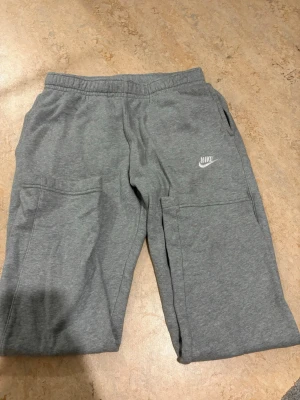 Grå mjukisbyxor från Nike M - Säljer ett par grå mjukisbyxor från Nike i storlek M. Byxorna har resår i midjan, sidofickor och en broderad Nike-logga på benet. Tillverkade i mjukt material med borstad insida för extra komfort. Perfekta för chill eller träning.