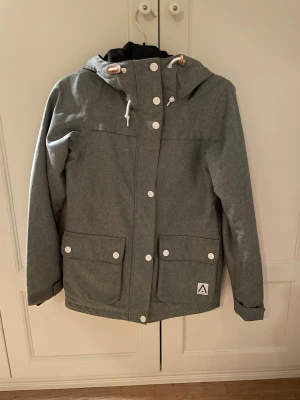 Grå jacka med huva från Wearcolour - Säljer en stilren grå jacka från Wearcolour med huva och stora fickor framtill. Jackan har vita knappar, dragkedja och snörning i huvan. Perfekt för dig som gillar en clean look och vill ha något funktionellt till vintern eller skidbacken.