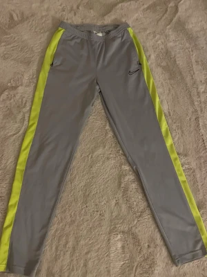 Grå Nike träningsbyxor med gula ränder - Säljer ett par grå träningsbyxor från Nike med gula sidopaneler längs benen. Byxorna har elastisk midja och smal passform, perfekta för sport eller chill. Tillverkade i ett mjukt och lätt syntetmaterial som andas bra. Klassisk Nike-logga på ena låret.