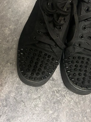 Svarta Louboutin sneakers med nitar - Säljer ett par svarta sneakers från Christian Louboutin med klassisk röd sula och coola svarta nitar på tån. Skorna har snörning, rund tå och är tillverkade i mocka och skinn. Perfekta för dig som vill sticka ut med en edgy och lyxig vibe.