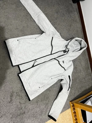 Ljusgrå vindjacka från Nike - Säljer en ljusgrå vindjacka från Nike med svarta detaljer och dragkedjor. Jackan har huva, långa ärmar och flera praktiska fickor, inklusive en på ärmen. Materialet är mjukt och känns sportigt, perfekt för dig som gillar en clean och enkel stil.