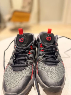 Nike Kevin Durant sneakers svart/röd - Säljer ett par Nike Kevin Durant sneakers i svart och grått med röda detaljer och vit swoosh. Skorna har snörning, lågt skaft och coolt mönstrad mesh på ovandelen. KD-logga på plösen och Kevin Durant-signatur på hälen. Perfekta för dig som gillar basketstil. Och det är storlek 43 och de är 27,5 cm 