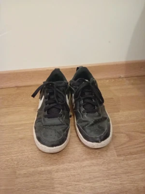 Svarta Nike sneakers med vit sula - Klassiska svarta Nike sneakers med vit sula och vit swoosh på sidan. Skorna har snörning och rund tå, samt en låg profil. Tillverkade i skinn och syntetmaterial. Perfekta för dig som gillar en sportig och enkel stil.