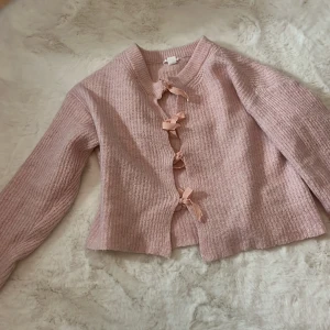 Rosa stickad kofta med rosetter - Supersöt rosa stickad kofta med långa ärmar och fyra dekorativa rosetter framtill istället för knappar. Perfekt för dig som gillar gulliga detaljer och mjuka material. Koftan har en croppad passform och bred ribbad kant vid halsen.