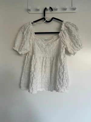 Vit puffärm blus från Gina Tricot - Supersöt vit blus från Gina Tricot med puffiga korta ärmar och vågig struktur i tyget. Blusen har en luftig passform och en lätt rynkad effekt som ger en romantisk vibe. Perfekt till jeans eller kjol för en fräsch look.