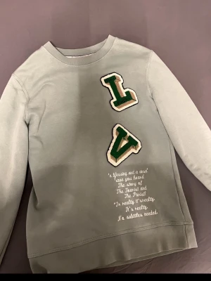 Louis Vuitton sweatshirt med tryck - Snygg grön sweatshirt från Louis Vuitton med stora gröna och vita LV-bokstäver framtill och coolt tryck med skelett och fågel på ryggen. Unik textdetalj på framsidan och oversized passform. Perfekt för dig som vill sticka ut med streetwear-vibe. Passar 15  - 17 som väger runt 70kg. 