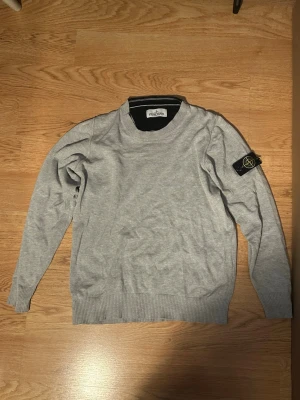 Grå Stone Island tröja junior - Säljer en grå långärmad tröja från Stone Island Junior med klassisk patch på ärmen. Tröjan har rund halsringning och ribbade muddar vid ärmslut och nederkant. Perfekt för dig som gillar streetwear och enkel stil.