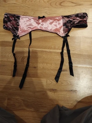 Rosa och svart satin strumpebandshållare - Säljer en snygg strumpebandshållare från H&M i rosa satin med svarta spetsdetaljer och svarta band. Den har små rosetter och justerbara band med knäppen. Perfekt för alla hjärtans dag  / Valentine's day. Jag har fler annonser uppe och ger mängd rabatt, så kolla gärna min sida!