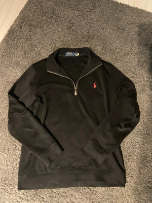 Svart half zip tröja Polo Ralph Lauren - Svart långärmad tröja från Polo Ralph Lauren med klassisk röd broderad logga på bröstet. Tröjan har en half zip-dragkedja vid halsen och är gjord i mjuk bomull. Perfekt för en clean och stilren look.
