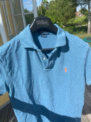 Blå pikétröja från Polo Ralph Lauren - Klassisk blå pikétröja från Polo Ralph Lauren med orange broderad logga på bröstet. Tröjan har korta ärmar, krage och två knappar framtill. Tillverkad i mjuk bomull som känns skön mot huden. Perfekt till sommaren för en clean och avslappnad look. OBS det finns ett litet hål i nacken