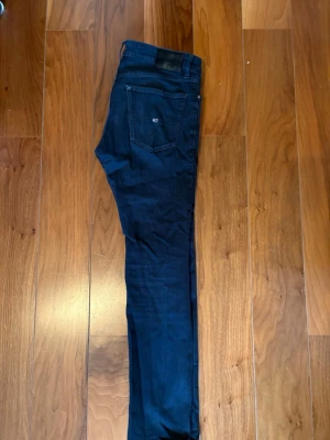  Tommy Jeans   - Säljer ett par mörkblå skinny jeans från Tommy Jeans i storlek 32. Jeansen har slitningar och hål på benen för en edgy look. Klassisk femficksmodell med låg midja och smal passform. Perfekta för dig som gillar streetstyle.