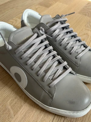 Gråa sneakers från Oliver Cabell - Snygga grå sneakers från Oliver Cabell med vita detaljer och rund tå. Skorna har klassisk snörning, vit sula och är tillverkade i slätt skinn. Insidan är fodrad i vitt tyg och hälen har en vit patch med guldfärgad logga. Perfekta för en clean och stilren look.