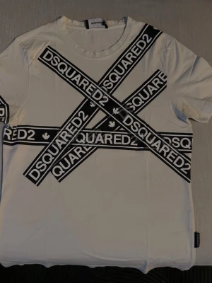 Vit DSQUARED2 t-shirt med logga - Säljer en vit t-shirt från DSQUARED2 i storlek S. T-shirten har svart tryck med stora DSQUARED2-loggor i kors över hela framsidan. Klassisk rund hals och korta ärmar. Tillverkad i mjuk bomull för skön känsla.