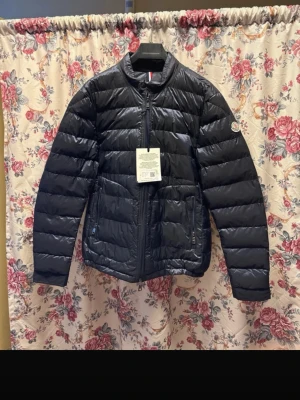 Svart Moncler pufferjacka - Säljer en svart pufferjacka från Moncler med klassisk quiltad design och rund hals. Jackan har dragkedja framtill, två sidofickor med dragkedja och Moncler-logga på ärmen. Materialet är lätt och glansigt, perfekt för kyliga dagar. NFC +