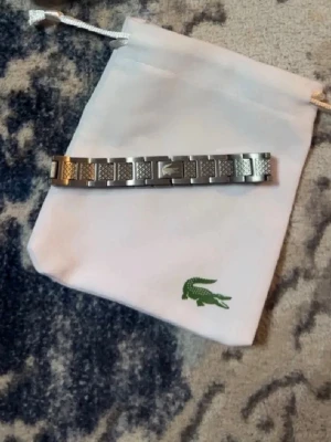 Stålarmband från Lacoste - Säljer ett stilrent armband i rostfritt stål från Lacoste. Armbandet har rektangulära länkar med graverade detaljer och den klassiska krokodilloggan på mitten. Perfekt accessoar för dig som gillar en clean och sportig look.