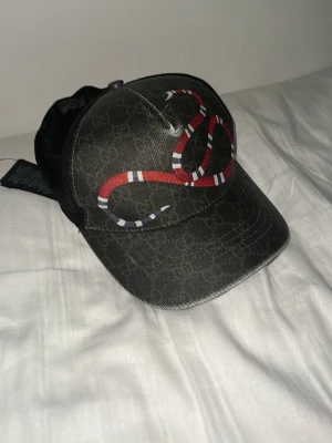 Gucci kingsnake print GG Supreme - Säljer en svart keps från Gucci med ikoniskt ormtryck i rött, vitt och blått på framsidan. Kepsen har diskret GG-mönster över hela materialet och mesh på sidorna och bak för extra ventilation. Justerbar passform och riktigt snygg streetwear-vibe. Den har storlek M men även 57 cm.