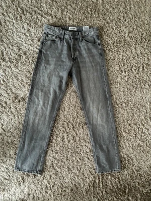 Grå jeans från Jack & Jones, straight fit - Säljer ett par gråa jeans från Jack & Jones i rak modell. Klassisk femficksdesign, normal midja och raka ben. Jeansen är tillverkade i bomull och har en snygg tvättad look. Perfekta till vardags för en avslappnad stil.