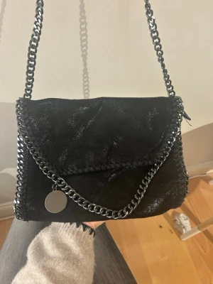 Svart axelväska med kedja - Cool svart axelväska med grov kedja i metall och snygga detaljer runt kanterna. Väskan har ett lock med magnetstängning och insidan är fodrad med leopardmönstrat tyg. Perfekt storlek för det viktigaste och har en rund metallbricka som dekoration.