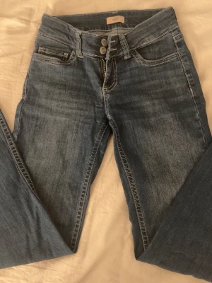 Blå jeans från Nelly med broderade fickor - Säljer ett par jättefina mellanblå jeans från Nelly med snygga broderade detaljer på bakfickorna. Jeansen har två knappar i midjan, klassisk femficksmodell och kontrastsömmar. Materialet är jeans och passformen är rak med låg midja.