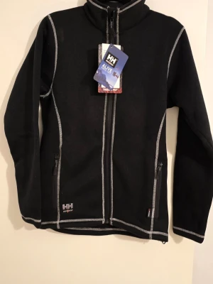 Svart fleecekofta Helly Hansen XS - Svart fleecekofta från Helly Hansen med vita kontrastsömmar och dragkedja framtill. Två fickor med dragkedja och hög krage. Polartec Thermal Pro-material som håller värmen. Snygg HH-logga på bröstet och ärmen. Perfekt för kyliga dagar.