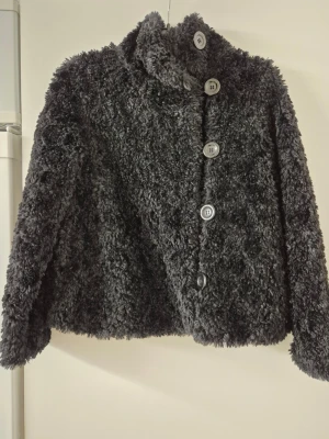 Teddyjacka - Mysig svart teddyjacka från Lindex med hög krage och stora knappar framtill. Jackan har en fluffig och mjuk känsla, perfekt för kyliga dagar. Enkel och stilren design som är lätt att matcha med olika outfits.