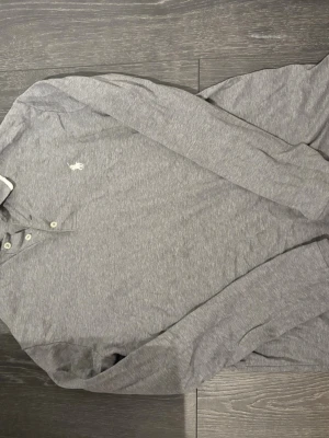 Grå långarmad piké från Polo Ralph Lauren - Snygg grå långärmad piké från Polo Ralph Lauren i slim fit. Skjortan har knappar vid halsen och den klassiska broderade loggan på bröstet. Tillverkad i mjuk bomull som känns skön mot huden.