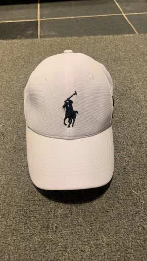 Vit keps från Polo Ralph Lauren - Säljer en stilren vit keps från Polo Ralph Lauren med klassisk svart polospelare broderad framtill och en stor svart 3:a på sidan. Justerbar baktill och ventilerande detaljer. Perfekt accessoar för en fin look.