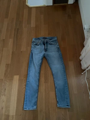 Ljusblå jeans från Nudie Jeans, 27/28 - Säljer ett par ljusblå jeans från Nudie Jeans i storlek 27/28. Jeansen har Nudie-loggan på bakfickorna och är tillverkade i slitstarkt bomullsmaterial. Modellen på jeansen är lean Dean 