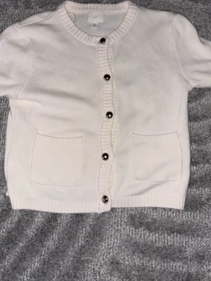 Vit kofta från H&M med knappar - Söt vit kofta från H&M med rund halsringning och korta ärmar. Tröjan har ribbade kanter, två framfickor och silverfärgade knappar framtill. Perfekt att slänga över en topp för en clean och enkel look.