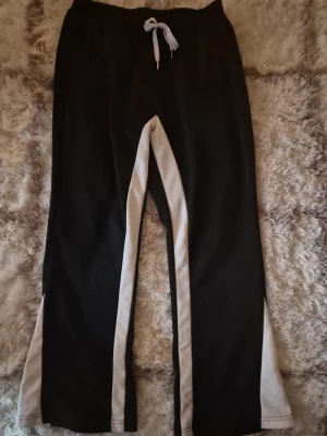 Svarta flared mjukisbyxor med vita detaljer - Säljer ett par svarta flared mjukisbyxor med vita sidopaneler och snörning i midjan. Byxorna har en loose passform och är perfekta för chill dagar eller träning. Materialet är mjukt och bekvämt, och designen är enkel men ändå snygg.