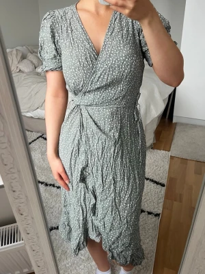 Prickig omlottklänning - Supersöt omlottklänning med vita prickar och puffiga korta ärmar. Klänningen har knyt i midjan, v-ringning och volangkant nertill. Okänt märke. Storlek S. Bra skick. Grön/grå färg <3