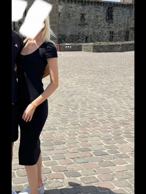 Svart ribbad knappklänning - Säljer en svart ribbad klänning med kort ärm och knappar framtill. Klänningen har en smickrande, kroppsnära passform och slutar under knät. Materialet är stretchigt och mjukt, perfekt för varma dagar. Enkel och stilren design som är lätt att matcha.