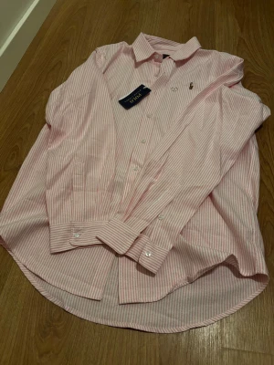 Ralph Lauren skjorta – oanvänd med tag! - Säljer en rosa/vit randig skjorta från Ralph Lauren (Polo). Helt oanvänd och fortfarande med tag kvar. Klassisk modell i hög kvalitet – funkar lika bra till vardag som till finare tillfällen.  Storlek: M Nypris: 1599 Mitt pris: 499  Kan skickas eller hämtas. Skicka DM vid intresse! 