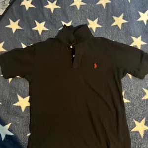 Svart pikétröja från Polo Ralph Lauren - Klassisk svart pikétröja från Polo Ralph Lauren i modellen Custom Slim Fit. Tröjan har korta ärmar, krage med knappar och den ikoniska röda logotypen broderad på bröstet. Tillverkad i mjuk bomull som känns skön mot huden.