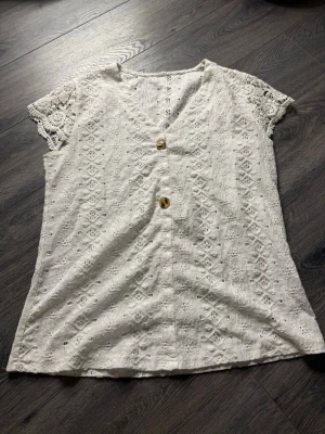 Vit spetsblus med knappar - Säljer en söt vit blus med v-ringning och två dekorativa knappar framtill. Blusen har korta, spetsiga ärmar och ett broderat spetsmönster över hela plagget. Perfekt för sommarens alla tillfällen och ger en romantisk vibe.