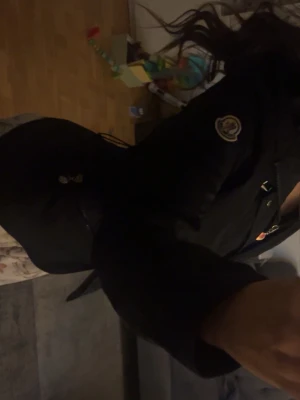 Svart vindjacka från Moncler med huva - Säljer en svart vindjacka från Moncler med huva och snygga detaljer som dragkedja och knappar framtill. Jackan har Moncler-logga på ärmen, justerbar midja och praktiska fickor med dragkedja. Perfekt för dig som vill ha en clean och stilren look.