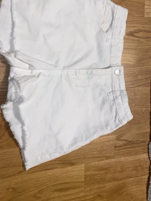 Vita jeansshorts från Lindex - Säljer ett par vita jeansshorts från Lindex i storlek 158. De har råa, fransiga kanter och klassisk femficksdesign. Perfekta till sommaren och har en clean look som passar till det mesta. Gjorda i bomull för skön känsla. Är midrise, bra skick använt några gånger. Säljer pga jag inte använder de längre, om du har några frågor meddela bara.✨