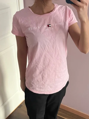 Rosa t-shirt från Tommy Hilfiger - Snygg ljusrosa t-shirt från Tommy Hilfiger i ekologisk bomull. Klassisk passform med korta ärmar och rund hals. Tryck med logga och text framtill samt liten flagga på ärmen. Perfekt för en chill och stilren look.