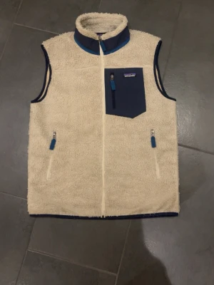 Beige fleeceväst från Patagonia  - Säljer en beige fleeceväst från Patagonia i storlek M. Västen har blå detaljer, hög krage och tre fickor med dragkedja, varav en bröstficka i kontrasterande blått tyg. Perfekt för lager-på-lager och outdoor-vibe.